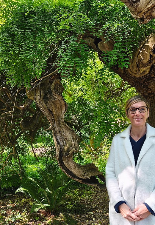 Barbara Masson en manteau clair, debout à côté d’un grand arbre noueux, dans une végétation luxuriante
