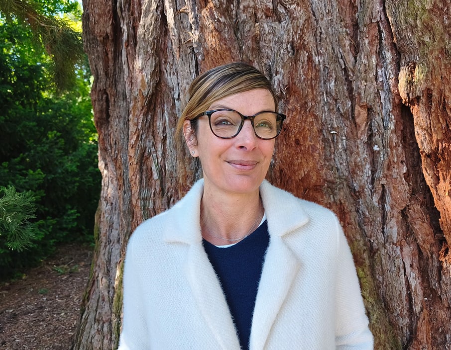 Barbara Masson, sophrologue et hypnothérapeute, en manteau clair devant l’écorce rouge d’un grand arbre
